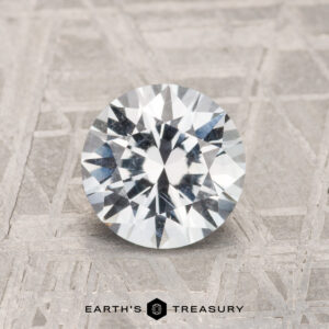 A 1.52-carat round brilliant-cut Montana Sapphire is shown on a textured gray surface with "EARTH'S TREASURY" displayed at the bottom.
