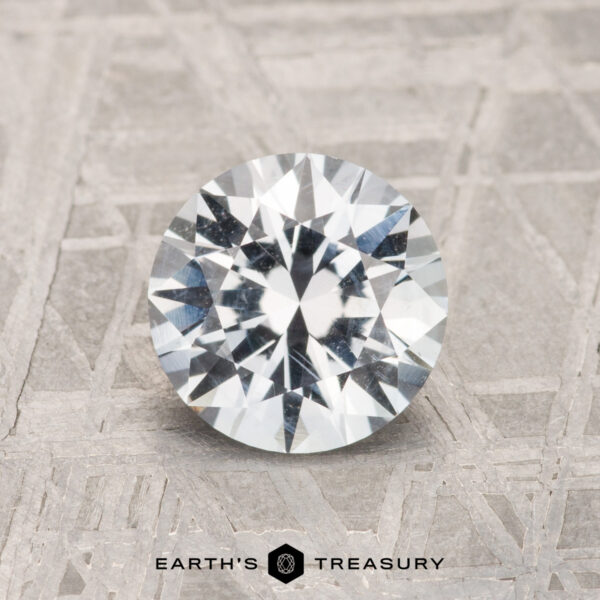 1.52-Carat Montana Sapphire A 1.52-carat round brilliant-cut Montana Sapphire is shown on a textured gray surface with "EARTH'S TREASURY" displayed at the bottom.