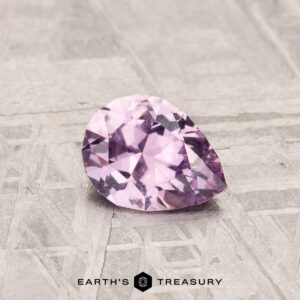 A pear-shaped, faceted 0.74-Carat Montana Sapphire in pale pink rests on a textured light gray surface, with "EARTH'S TREASURY" displayed at the bottom.
