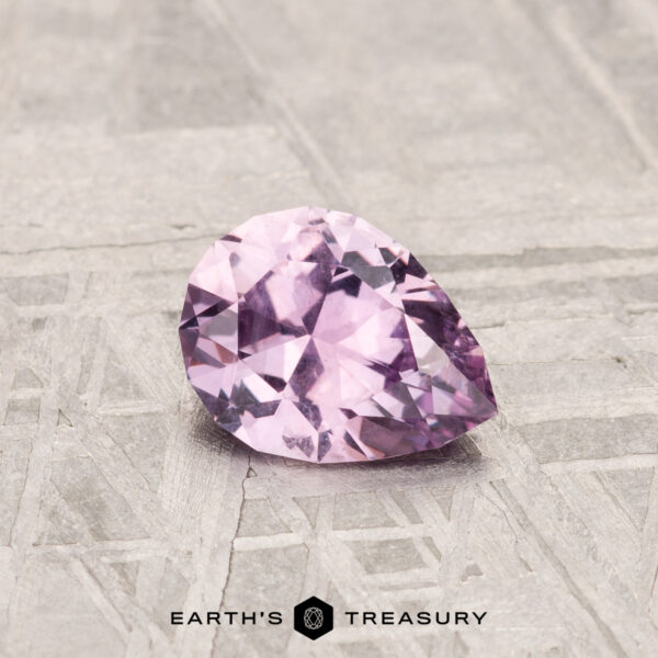 0.74-Carat Montana Sapphire A pear-shaped, faceted 0.74-Carat Montana Sapphire in pale pink rests on a textured light gray surface, with "EARTH'S TREASURY" displayed at the bottom.