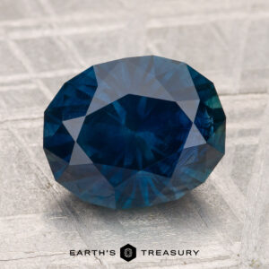 A 3.80-carat heated Montana sapphire in faceted oval blue rests on a textured gray surface, with "Earth's Treasury" at the bottom of the image.