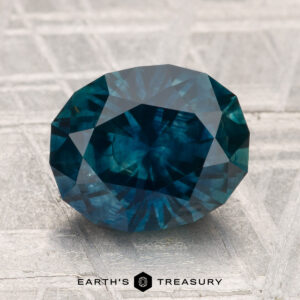 A 3.52-carat heated Montana sapphire, cut in an oval shape and displaying a deep blue color, rests on a gray textured surface with "EARTH'S TREASURY" text below.