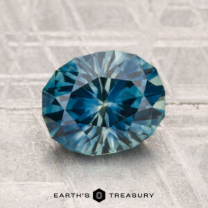 A 2.37-carat oval-cut blue-green Montana sapphire (heated) with angular facets rests on a textured gray surface, featuring the "EARTH'S TREASURY" logo and text at the bottom.