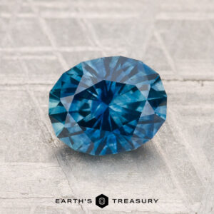 A 1.74-carat heated Montana sapphire, cut in a faceted oval shape, is shown on a textured gray surface with "EARTH'S TREASURY" at the bottom.