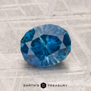 A 1.64-carat heated Montana sapphire with a faceted blue appearance rests on a light, textured surface, displaying "EARTH'S TREASURY" at the bottom.