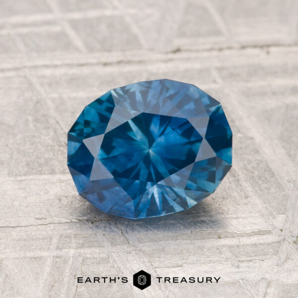 1.64-Carat Montana Sapphire (Heated) A 1.64-carat heated Montana sapphire with a faceted blue appearance rests on a light, textured surface, displaying "EARTH'S TREASURY" at the bottom.