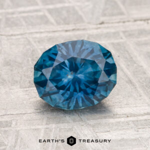 A 1.57-carat heated Montana sapphire is displayed on a textured gray surface with the text "EARTH'S TREASURY" below it.