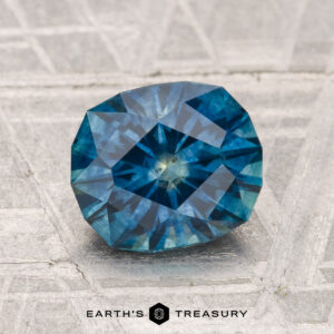 A 2.02-carat heated Montana sapphire is displayed on a textured gray surface with "EARTH'S TREASURY" at the bottom.
