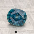 A faceted oval blue-green 2.00-carat heated Montana sapphire rests on a textured light gray surface, with "EARTH'S TREASURY" visible at the bottom.