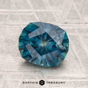 A faceted oval blue-green 2.00-carat heated Montana sapphire rests on a textured light gray surface, with "EARTH'S TREASURY" visible at the bottom.