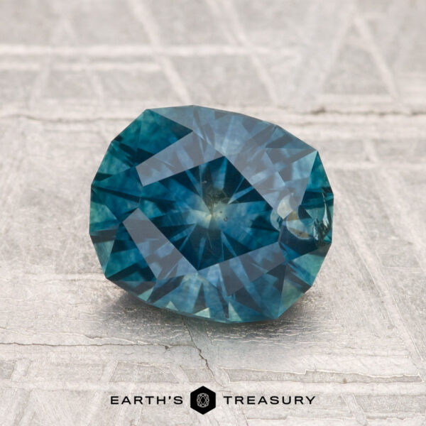 2.00-Carat Montana Sapphire (Heated) A faceted oval blue-green 2.00-carat heated Montana sapphire rests on a textured light gray surface, with "EARTH'S TREASURY" visible at the bottom.