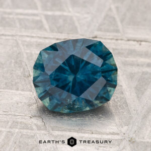 A 1.98-carat heated Montana sapphire with blue-green facets is displayed on a textured gray surface, with "EARTH'S TREASURY" text at the bottom.