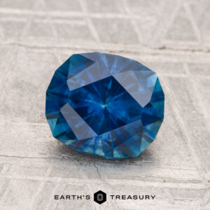A 1.91-carat heated Montana sapphire with an oval cut is displayed on a textured light gray surface, featuring the text "Earth's Treasury" at the bottom.