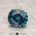 A 1.90-carat heated Montana sapphire gemstone with blue-green hues is displayed on a textured gray surface, featuring the text "EARTH'S TREASURY" at the bottom.