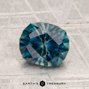 A 1.90-carat heated Montana sapphire gemstone with blue-green hues is displayed on a textured gray surface, featuring the text "EARTH'S TREASURY" at the bottom.