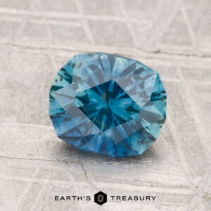 A 1.80-carat heated Montana sapphire with visible dark inclusions rests on a textured gray surface. "EARTH'S TREASURY" is labeled at the bottom.