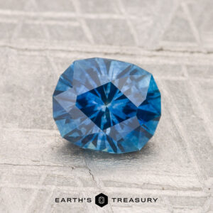 A 1.34-carat heated Montana sapphire rests on a textured surface, with the "Earth's Treasury" logo and text visible at the bottom of the image.