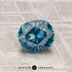 A 1.23-carat heated Montana sapphire with a geometric faceted cut is showcased on a textured gray surface, featuring the "Earth's Treasury" logo at the bottom.