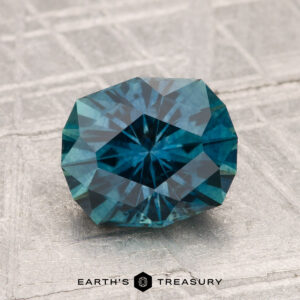A 2.24-carat heated Montana sapphire with a geometric cut rests on a textured gray surface. The Earth's Treasury logo is displayed at the bottom of the image.