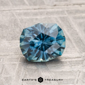 A faceted, round 1.64-carat heated Montana sapphire in teal-blue rests on a textured gray surface with "EARTH'S TREASURY" below.
