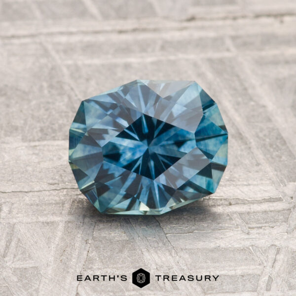 1.64-Carat Montana Sapphire (Heated) A faceted, round 1.64-carat heated Montana sapphire in teal-blue rests on a textured gray surface with "EARTH'S TREASURY" below.