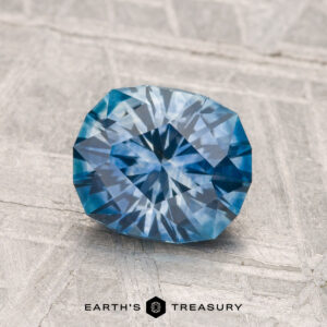 A 1.62-carat cushion-cut heated Montana sapphire is displayed on a light textured surface with "EARTH'S TREASURY" at the bottom.