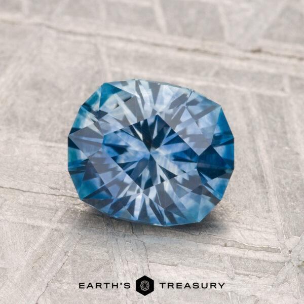 A 1.62-carat cushion-cut heated Montana sapphire is displayed on a light textured surface with "EARTH'S TREASURY" at the bottom.
