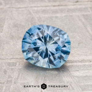 A 1.51-carat heated Montana sapphire with a cushion cut is displayed on a textured gray surface. The Earth's Treasury logo appears at the bottom.