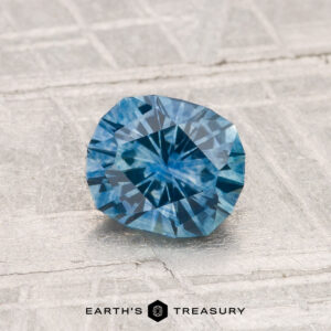 A faceted blue 1.07-carat heated Montana sapphire with a starburst pattern is displayed on a light textured surface above the text "Earth's Treasury.