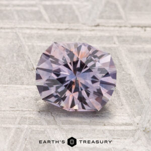 A round, faceted 1.41-carat Montana sapphire (heated) with a light lavender hue rests on a textured, light-colored surface. "Earth's Treasury" is displayed at the bottom of the image.