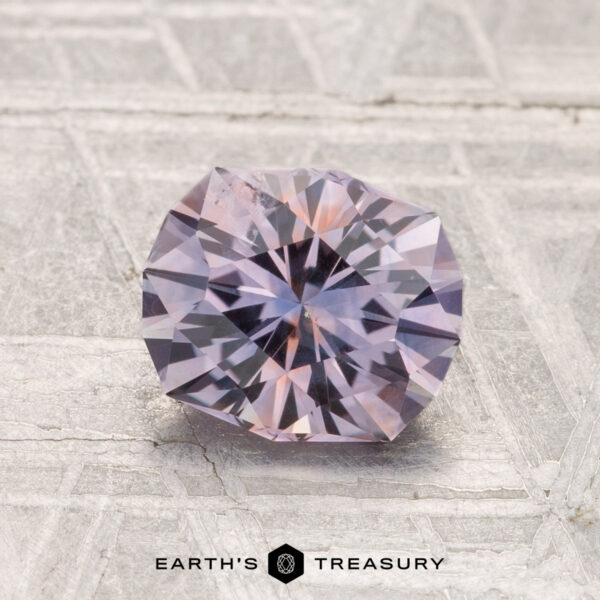 1.41-Carat Montana Sapphire (Heated) A round, faceted 1.41-carat Montana sapphire (heated) with a light lavender hue rests on a textured, light-colored surface. "Earth's Treasury" is displayed at the bottom of the image.