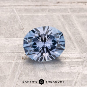 A 1.07-carat heated Montana sapphire, cut in a round brilliant style, is shown on a textured light gray surface with "EARTH'S TREASURY" at the bottom.