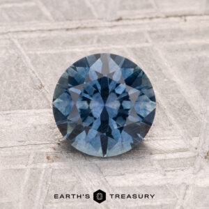 A 1.33-carat round-cut heated Montana sapphire is displayed on a textured gray metallic surface with the Earth’s Treasury logo at the bottom.