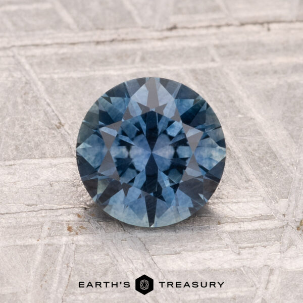 1.33-Carat Montana Sapphire (Heated) A 1.33-carat round-cut heated Montana sapphire is displayed on a textured gray metallic surface with the Earth’s Treasury logo at the bottom.