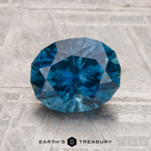 A 2.21-carat heated Montana sapphire, faceted in an oval shape, rests on a textured gray background with the "Earth's Treasury" logo at the bottom.