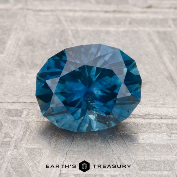2.21-Carat Montana Sapphire (Heated) A 2.21-carat heated Montana sapphire, faceted in an oval shape, rests on a textured gray background with the "Earth's Treasury" logo at the bottom.
