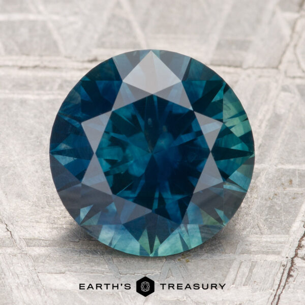 4.12-Carat Montana Sapphire (Heated) A 4.12-carat heated Montana sapphire, round and faceted in blue-green hues, is displayed on a textured metallic surface with "Earth's Treasury" below.