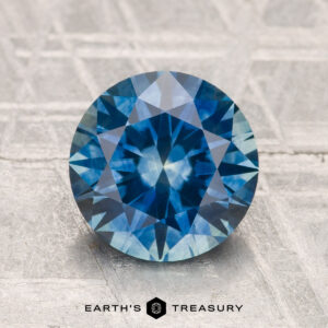 A 2.53-carat heated Montana sapphire, round brilliant-cut and blue, is displayed on a textured surface with the text "EARTH'S TREASURY" below it.