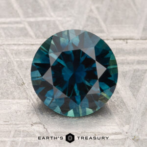 A 2.30-carat heated Montana Sapphire gemstone, round and blue-green with facets, is displayed on a textured gray surface with "EARTH'S TREASURY" below.
