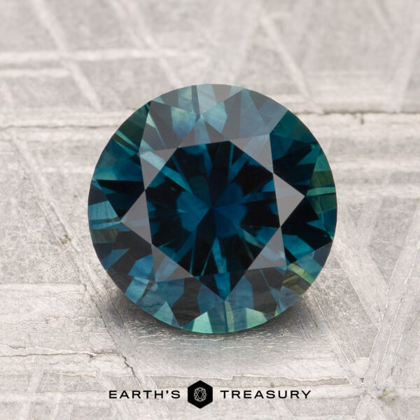 2.30-Carat Montana Sapphire (Heated) A 2.30-carat heated Montana Sapphire gemstone, round and blue-green with facets, is displayed on a textured gray surface with "EARTH'S TREASURY" below.