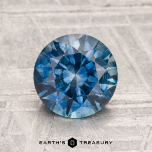 A round, faceted blue 1.89-carat Montana Sapphire (Heated) is displayed on a textured surface, with "Earth's Treasury" written below.