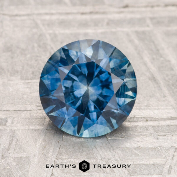1.89-Carat Montana Sapphire (Heated) A round, faceted blue 1.89-carat Montana Sapphire (Heated) is displayed on a textured surface, with "Earth's Treasury" written below.