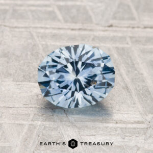 A 1.28-carat, round brilliant-cut heated Montana sapphire in light blue is displayed on a textured gray surface with "Earth's Treasury" below.