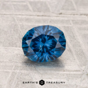 A 1.23-carat heated Montana sapphire, round brilliant-cut and blue, is shown on a textured light gray surface with "Earth's Treasury" below.