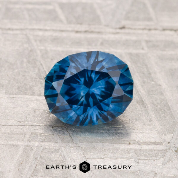 1.23-Carat Montana Sapphire (Heated) A 1.23-carat heated Montana sapphire, round brilliant-cut and blue, is shown on a textured light gray surface with "Earth's Treasury" below.