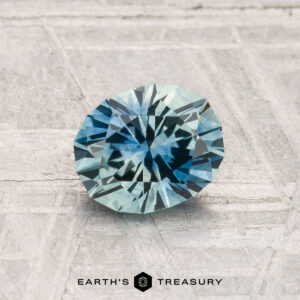 A 1.21-carat oval-cut heated Montana sapphire is displayed on a textured gray surface with the "Earth's Treasury" logo and text beneath it.