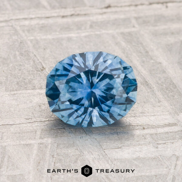 1.18-Carat Montana Sapphire (Heated) A 1.18-carat heated Montana sapphire, round and faceted, is displayed on a textured light gray surface with "EARTH'S TREASURY" at the bottom of the image.