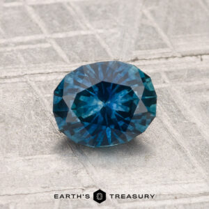 A 1.07-carat heated Montana sapphire with round, faceted cuts is shown on a textured gray surface, featuring "EARTH'S TREASURY" at the bottom.