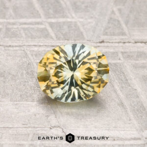 A 1.03-carat heated Montana sapphire with yellow and green hues rests on a textured gray surface. "EARTH'S TREASURY" is displayed at the bottom of the image.