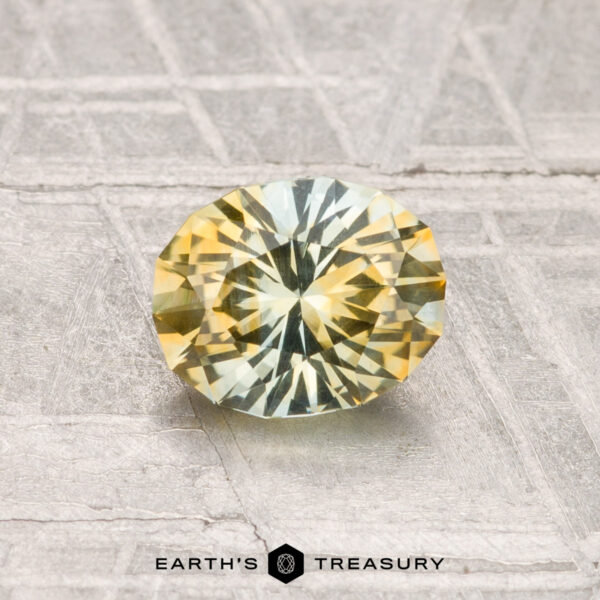 1.03-Carat Montana Sapphire (Heated) A 1.03-carat heated Montana sapphire with yellow and green hues rests on a textured gray surface. "EARTH'S TREASURY" is displayed at the bottom of the image.